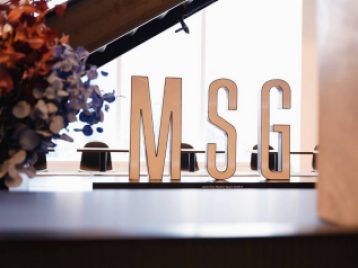 Madison Square Garden MSG logo sign inside with some flowers to the left in the foreground, and a staircase and windows framing it in the background. 